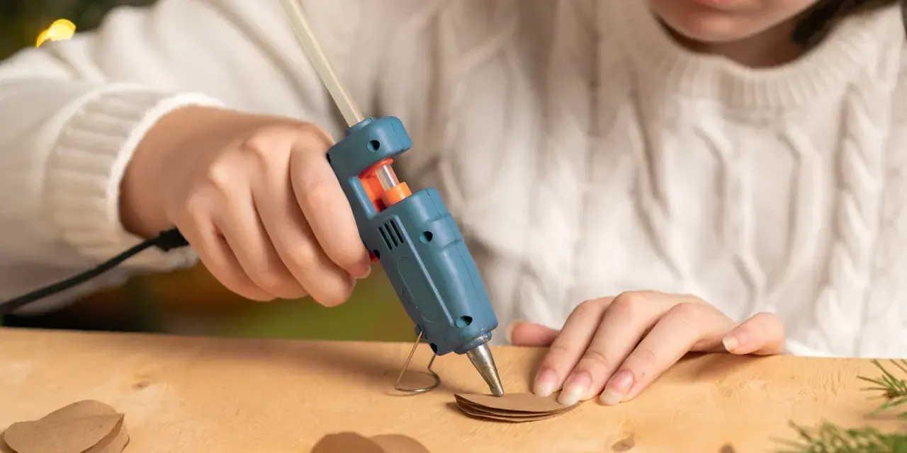 A child uses a hot glue gun to craft heart shapes from brown paper, wondering if hot glue holds up in water for their project.