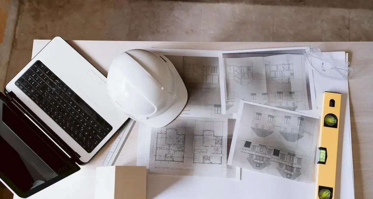 Laptop, kask, poziomica i plany architektoniczne – kompletny zestaw do pracy nad projektem technicznym.