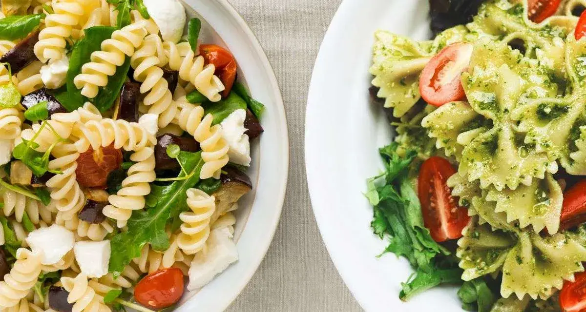 Due ciotole di pasta fredda gourmet: una con fusilli, melanzane, pomodorini e mozzarella; l'altra con farfalle al pesto e pomodorini.