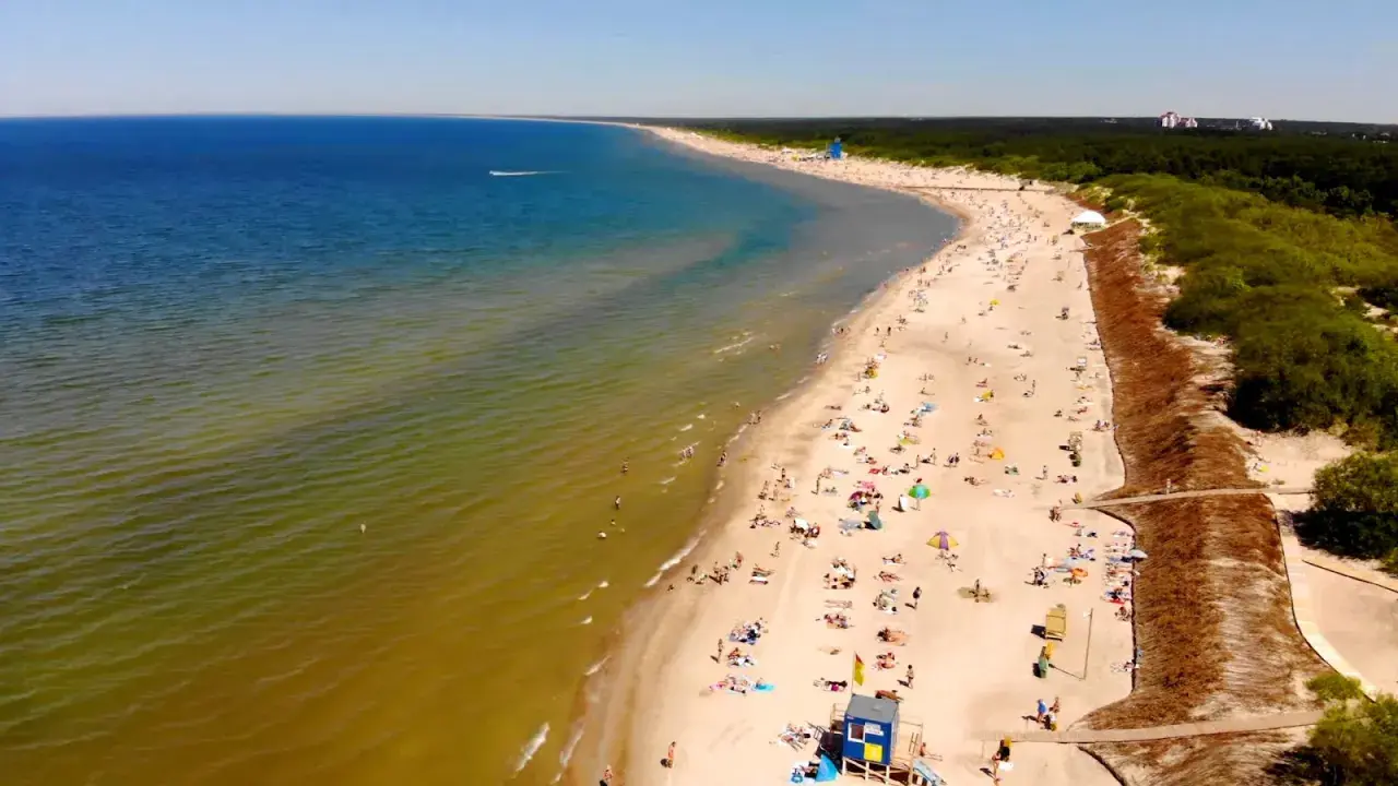 Słoneczna plaża na Litwie, gdzie turkusowe morze łagodnie przechodzi w zielonkawe wody. Tłumy plażowiczów odpoczywają na piasku, ciesząc się letnim dniem.