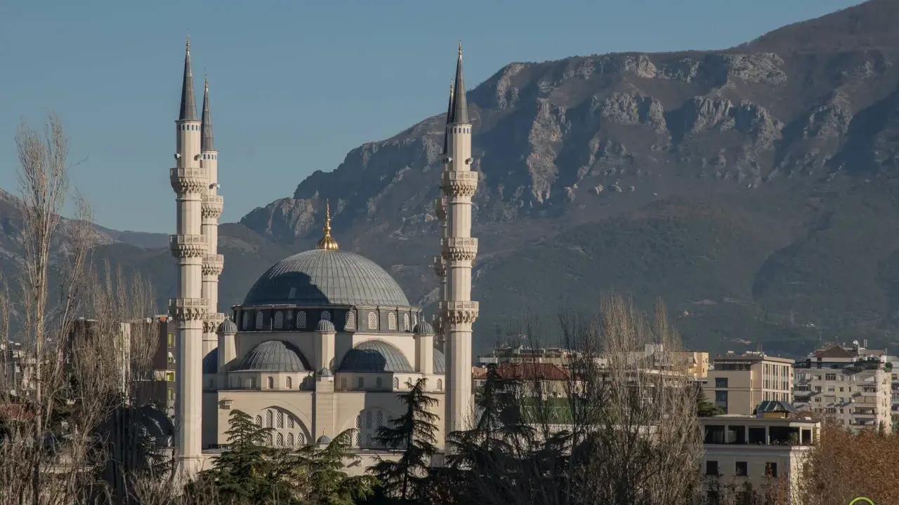 Imponujący meczet z minaretami, górujący nad budynkami stolicy Albanii, na tle skalistych gór.