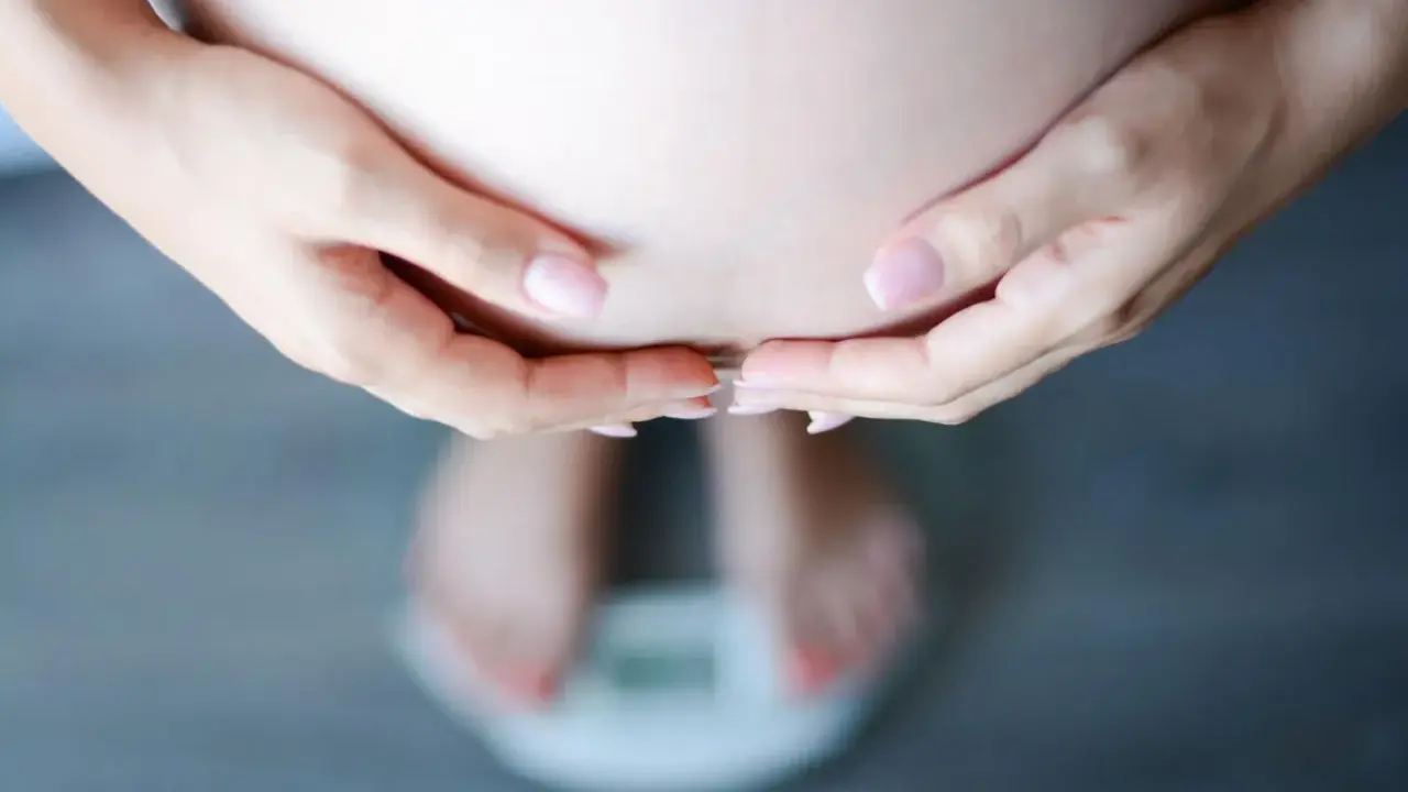 Dłonie obejmujące brzuch ciężarnej, stojącej na wadze. Schudłam w ciąży 10 kg, co widać na wadze.