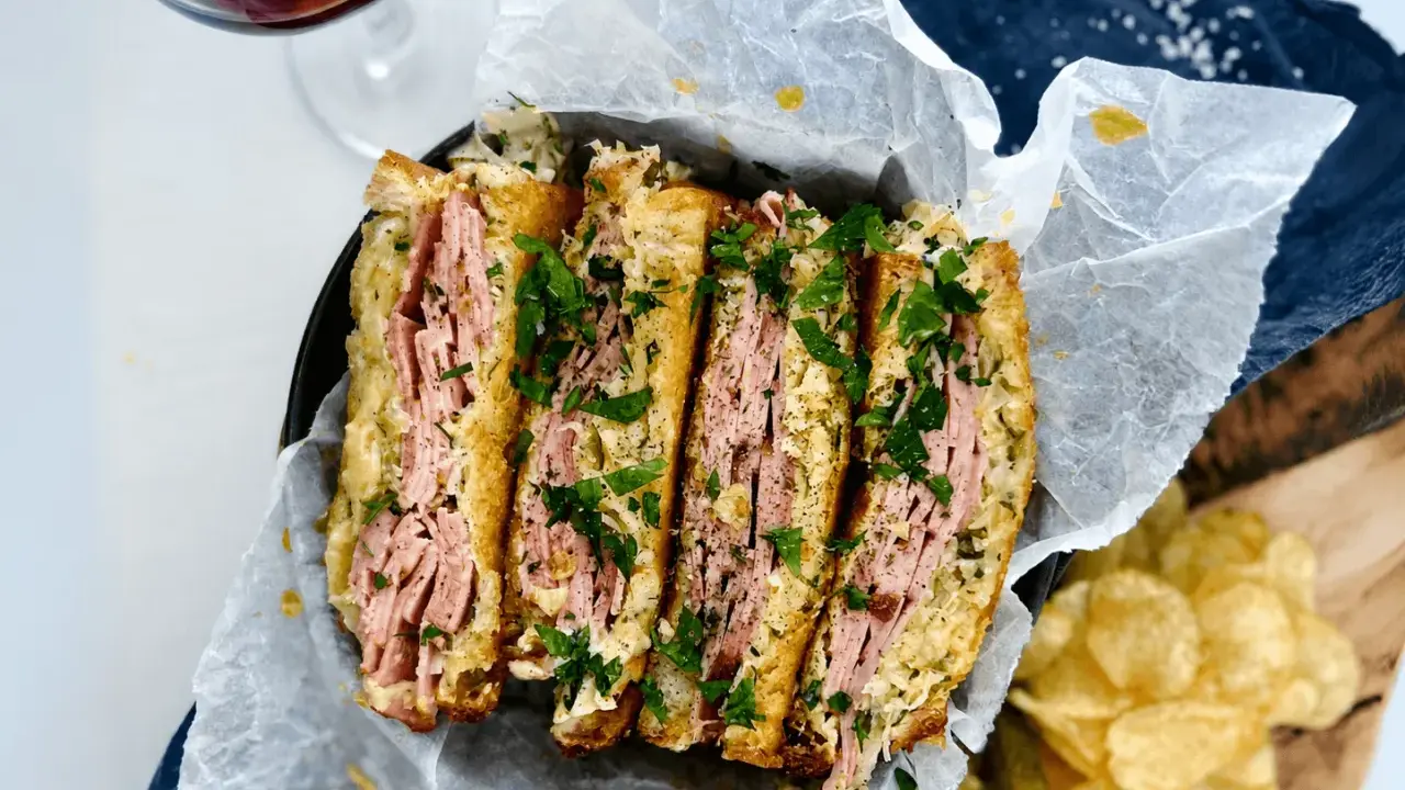 En saftig reuben sandwich, skuren i fyra bitar och toppad med färsk persilja. Serveras med krispiga potatischips och en glas rödvin.