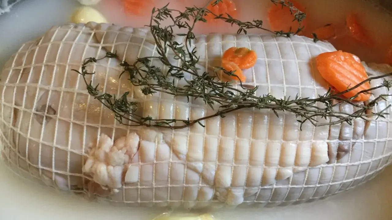Préparation de la cuisson tête de veau cocotte. Le veau est ficelé, entouré de carottes et de thym dans un bouillon.