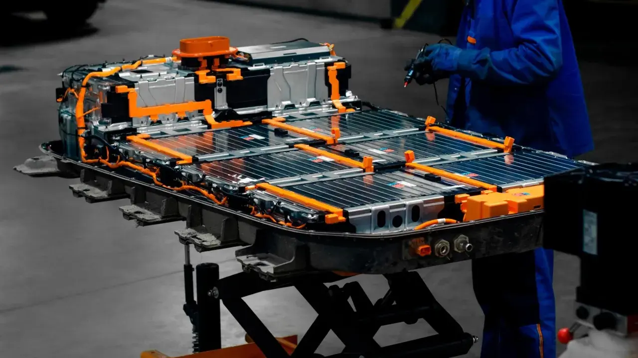 Assemblaggio di batterie per macchine elettriche, con cavi arancioni e celle metalliche. Un tecnico in tuta blu lavora sul pacco batteria.