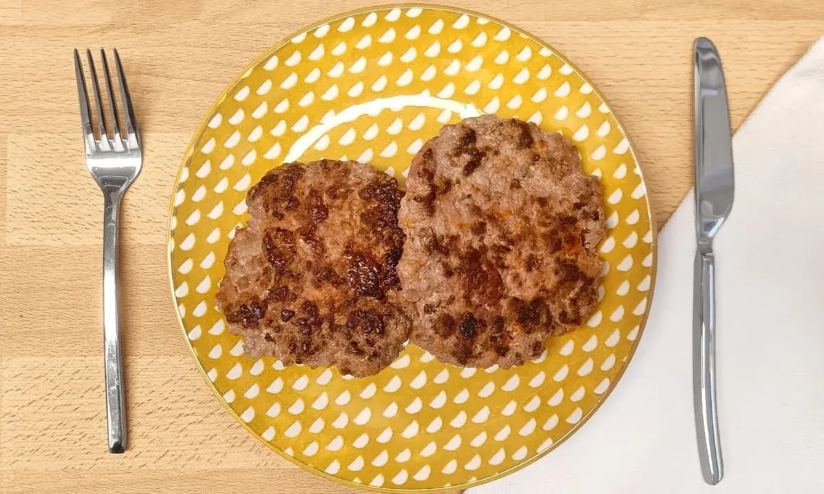 Dos hamburguesas para bebes, doradas y jugosas, servidas en un plato amarillo con puntos blancos. Cubiertos a los lados.