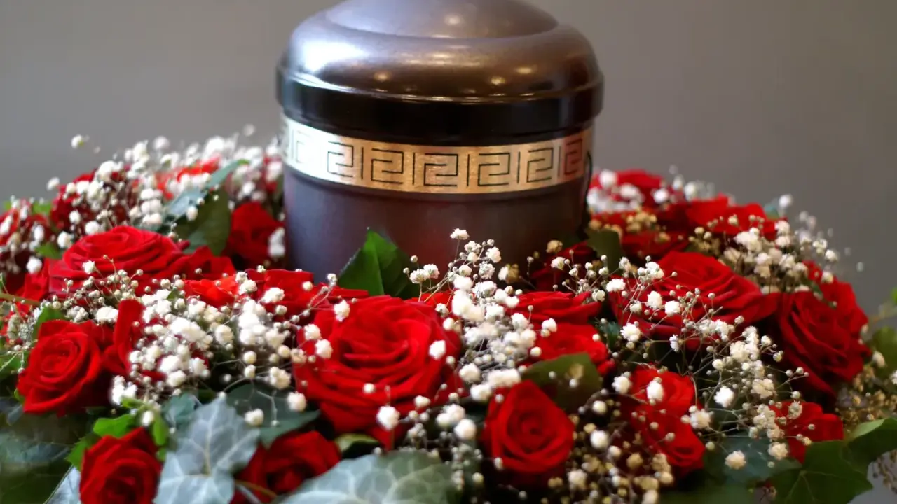 Ciemna urna pogrzebowa otoczona wieńcem z czerwonych róż i białych drobnych kwiatów.