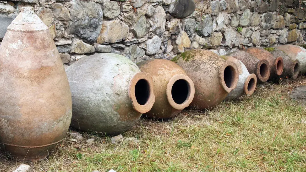 Antiguas ánforas de vino yacen en la hierba, algunas cubiertas de musgo, contra un muro de piedra.