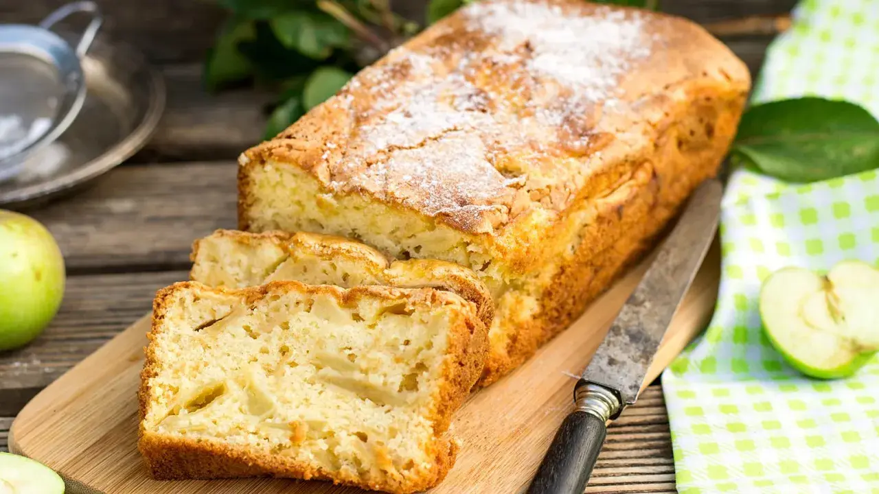 Szybkie ciasto z jabłkami, posypane cukrem pudrem, pokrojone na kromki, leży na desce obok jabłek i noża.