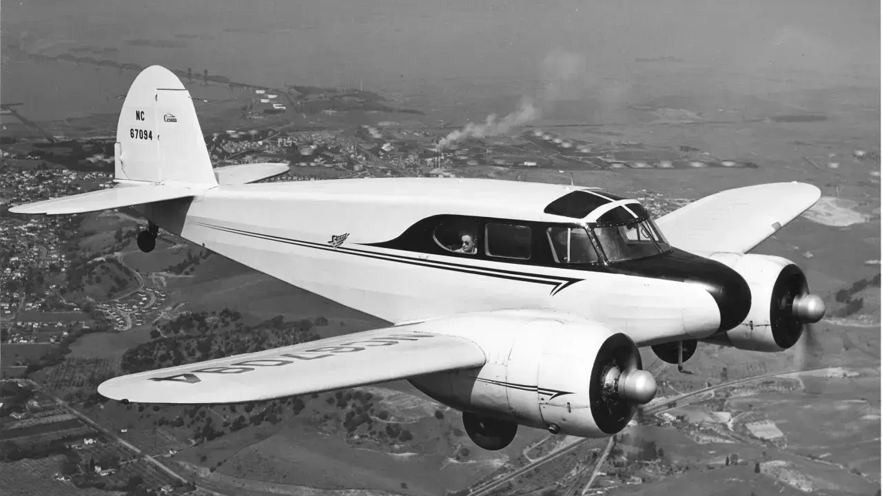 Ein weißes, zweimotoriges hochdecker flugzeug fliegt über eine Stadt und Industrieanlagen.