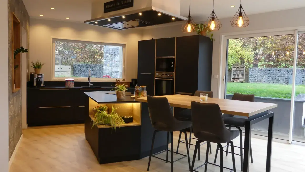 Cuisine noir et bois, mur gris texturé. Îlot central avec éclairage, table haute et tabourets. Fenêtre donnant sur jardin.