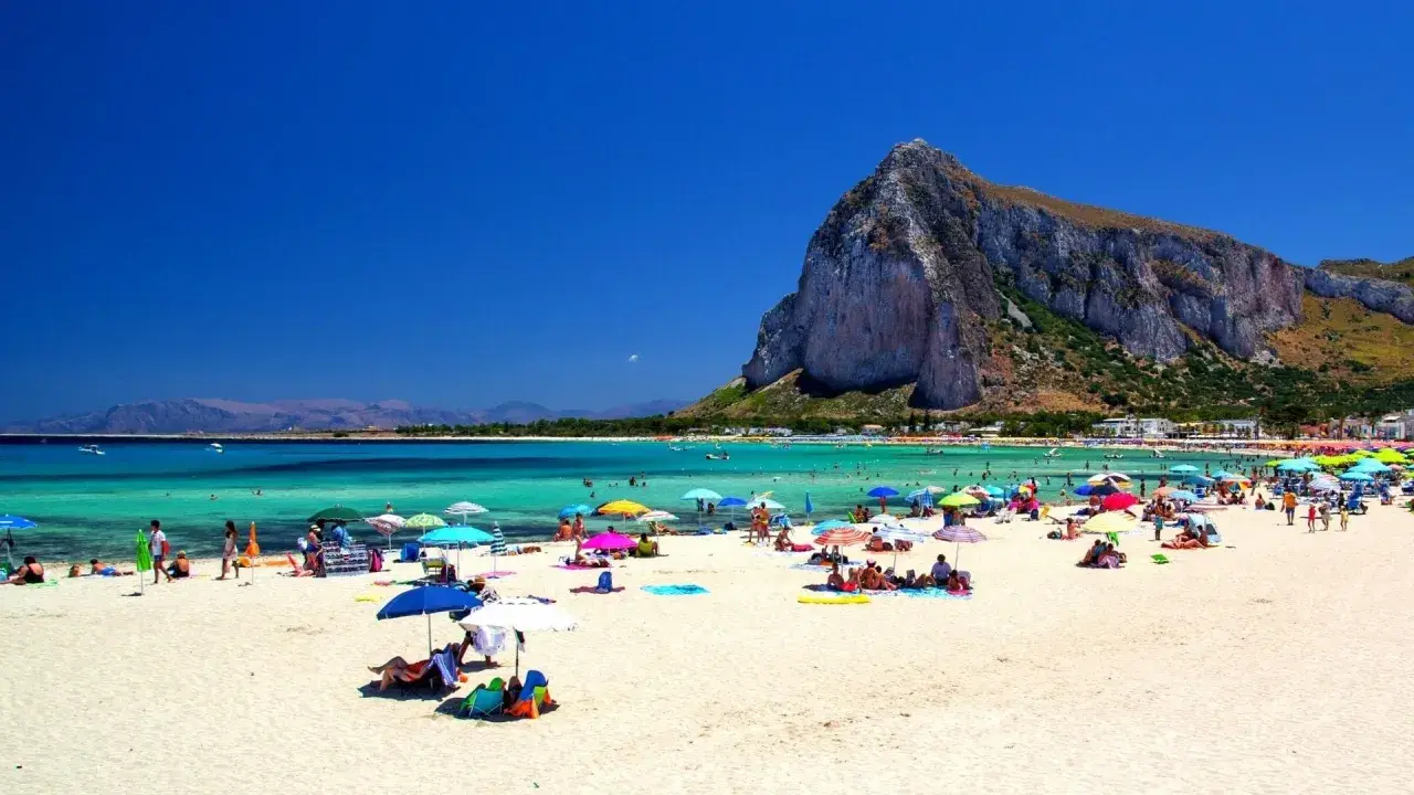 Spiaggia affollata con ombrelloni colorati, gente che si gode le vacanze al mare con bambini, acque turchesi e una montagna imponente sullo sfondo.