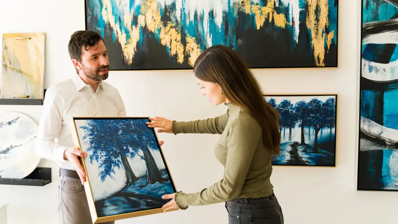 A man and woman admire a painting of blue trees at an art exhibition.