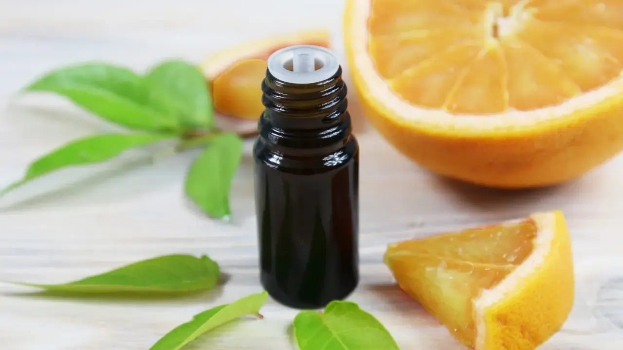 Flacon d'huile essentielle de citrus sinensis, entouré de feuilles vertes et de morceaux d'orange fraîche sur fond de bois clair.
