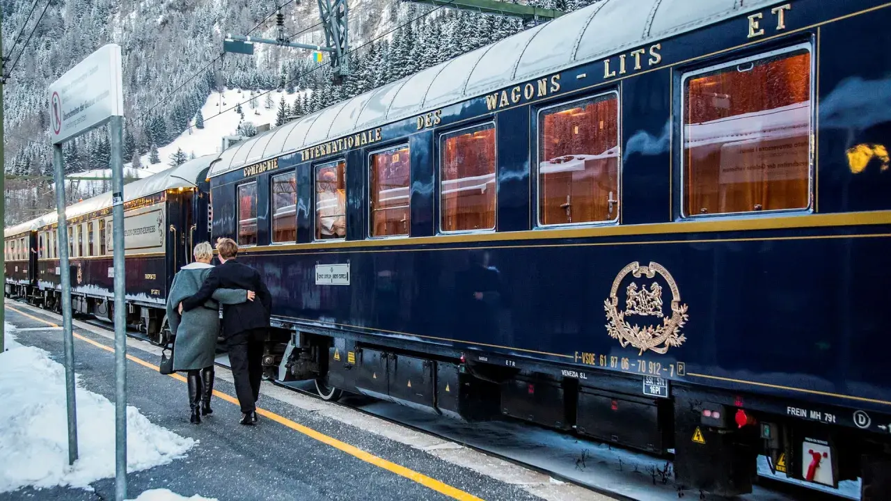 Ett par omfamnar varandra vid en snötäckt station, redo för en resa med Orientexpressen. Tågets mörkblå vagnar med gyllene detaljer vittnar om dess klassiska orientexpressen rutt.