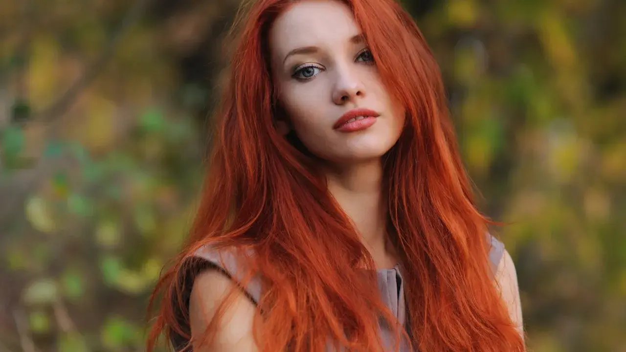 Eine Frau mit leuchtend roten Haaren steht im Freien. Ihre langen, feurigen Haare umrahmen ihr Gesicht, während sie in die Kamera blickt. Die Natur im Hintergrund ist unscharf und zeigt herbstliche Farben.