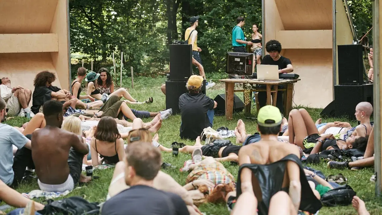 Un gruppo di persone rilassate su un prato in un **boutique festival**, con un DJ che suona musica in sottofondo.