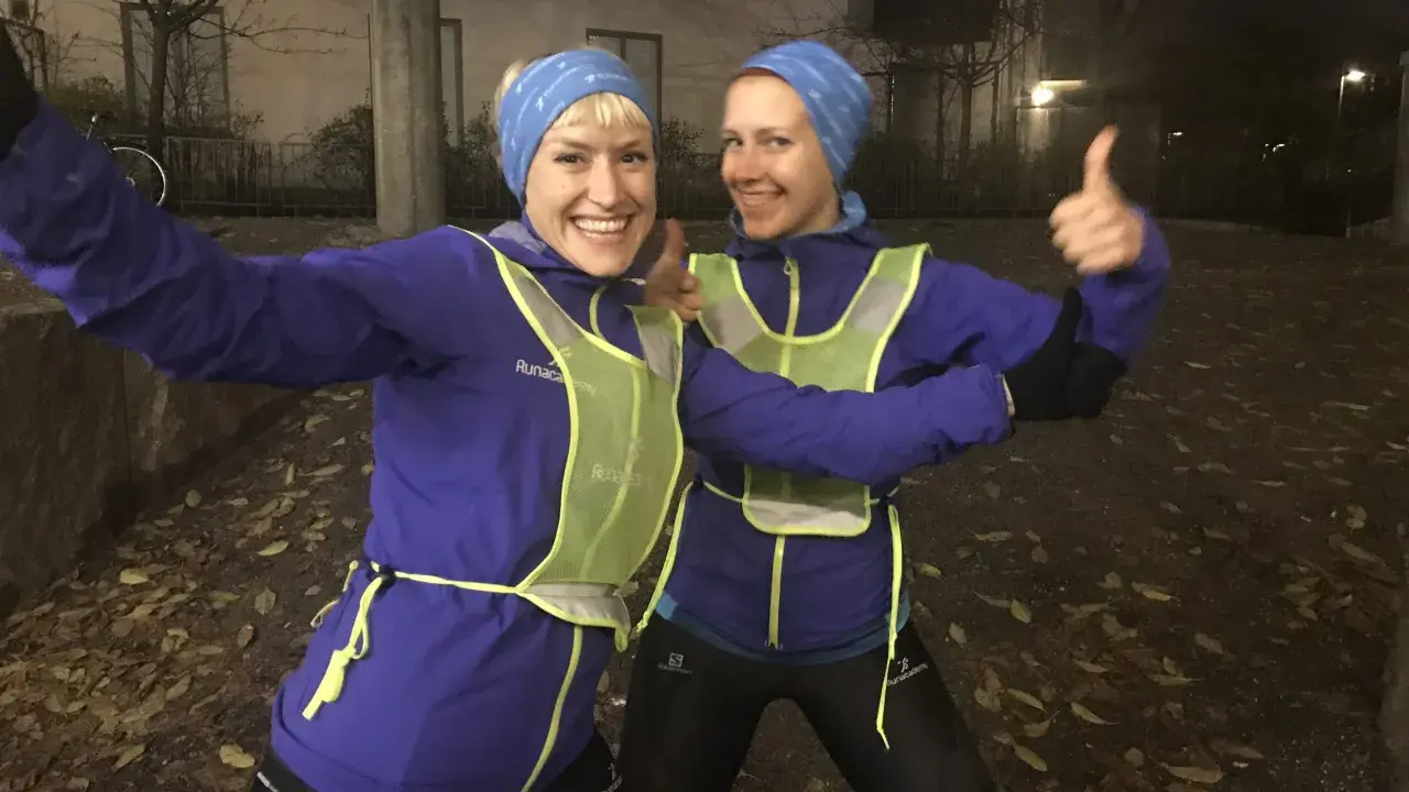 Två glada löpare i blå jackor och reflexvästar ger tummen upp, redo för ett maraton.