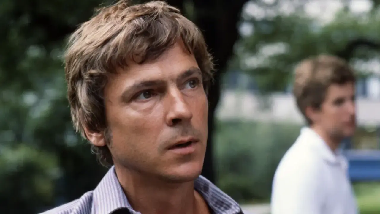 Carl Ivar Nilsson, en man med rufsigt hår och en randig skjorta, tittar bort. En annan man syns suddigt i bakgrunden.