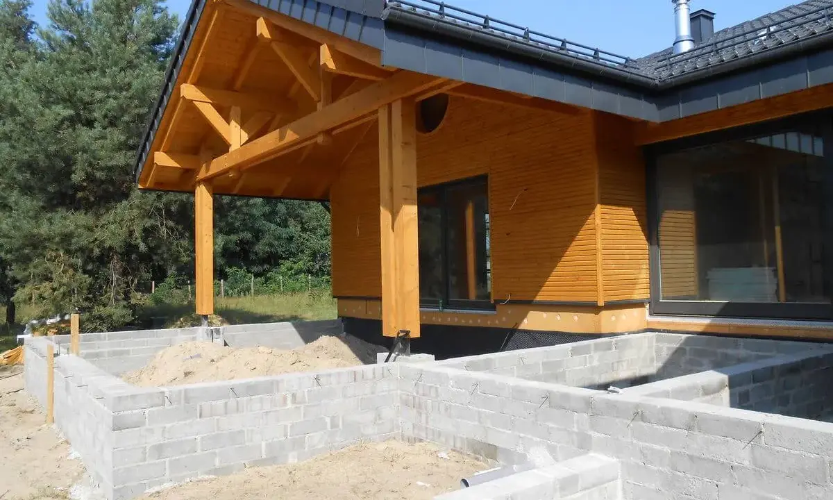 Drewniany dom w budowie, z widocznym fundamentem z bloczków betonowych i dachem o kącie nachylenia 12 stopni.
