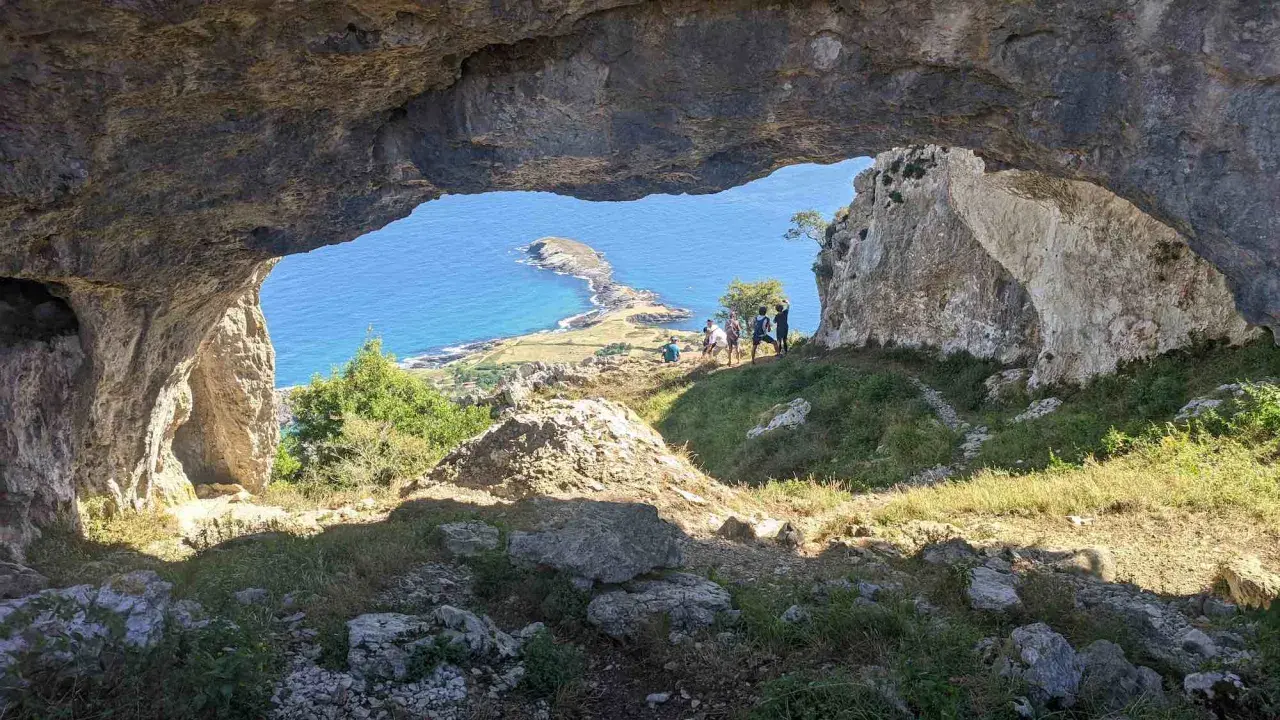 Vista desde el "ojo del diablo", una cueva natural, hacia un paisaje costero con un istmo y el mar azul. Un grupo de personas observa el panorama.