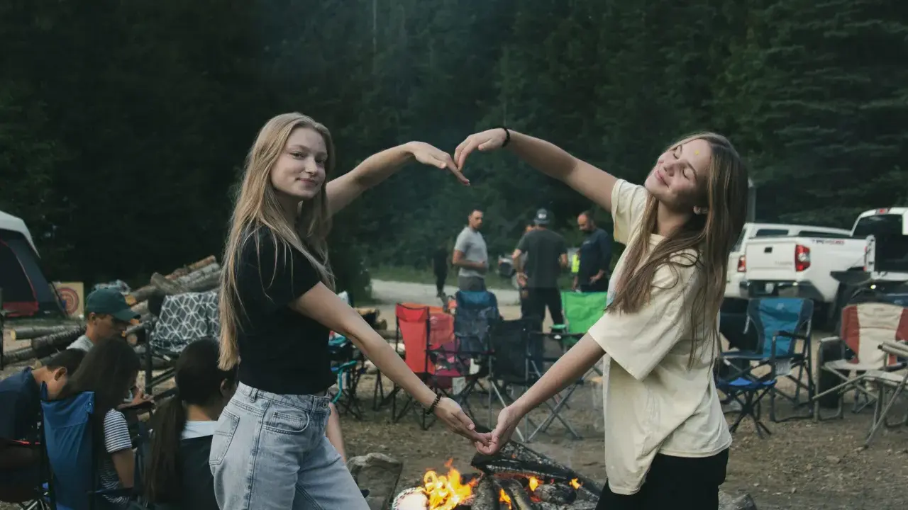 Dwie dziewczyny tworzą serce z dłoni na tle ogniska i lasu. To idealny okres na wakacjach, pełen radości i wspólnych chwil.