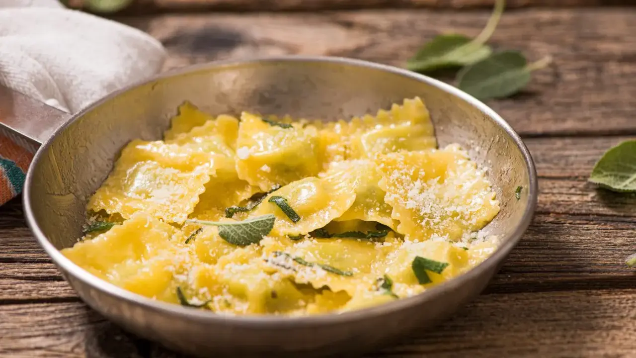 Ravioli ricotta e spinaci dorati, conditi con parmigiano e foglie di salvia fresca, serviti in una ciotola metallica su un tavolo di legno rustico.