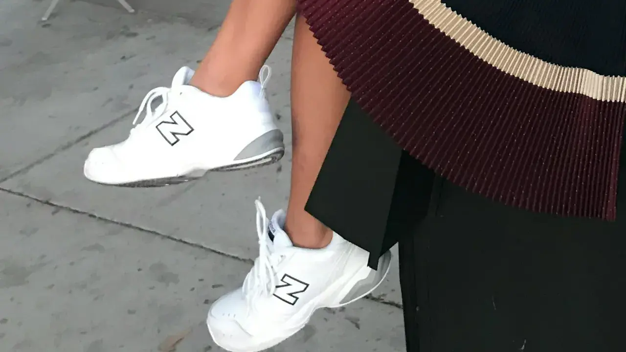 White New Balance dad shoes with a black "N" logo, paired with a pleated skirt.