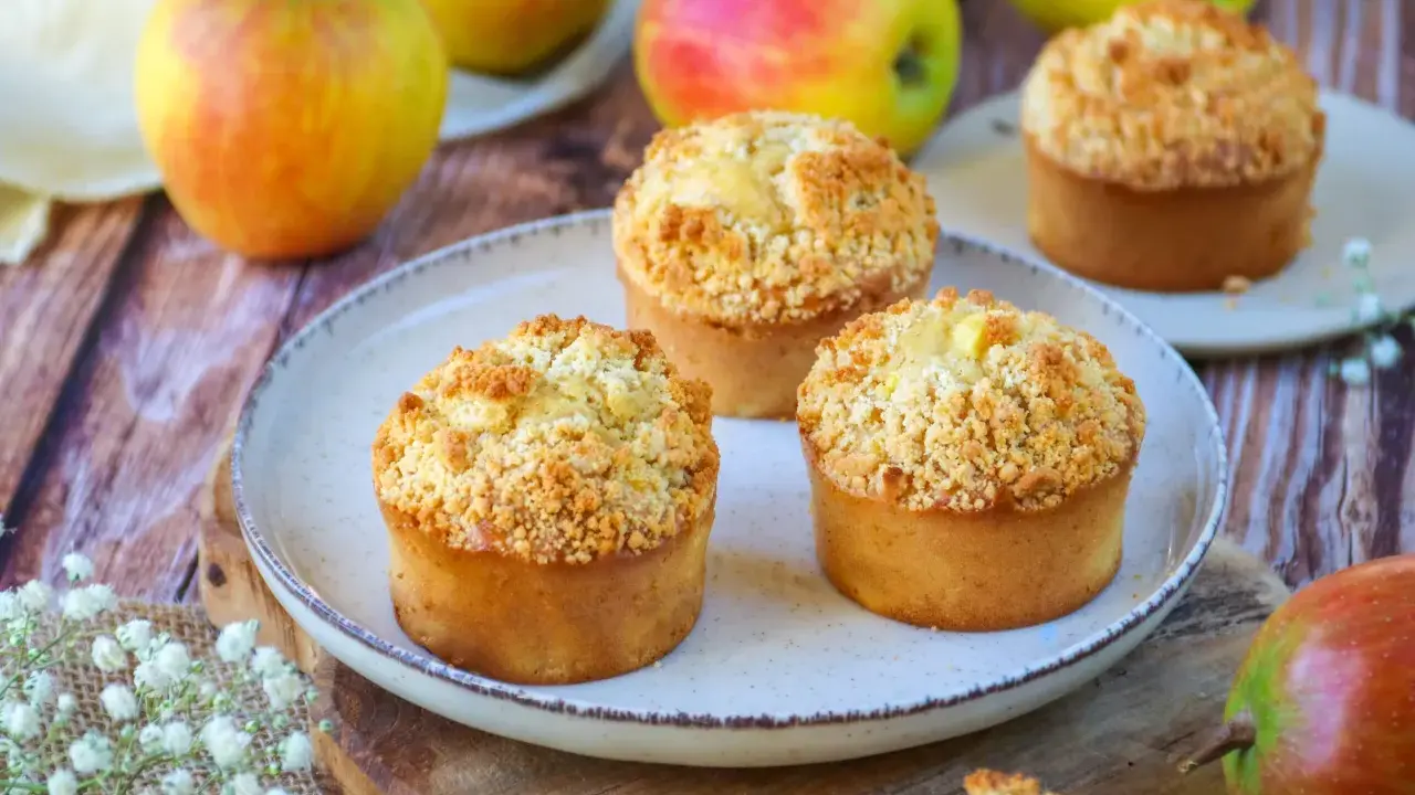 Trois muffins pomme amande dorés avec un crumble croustillant, entourés de pommes fraîches sur une table en bois.