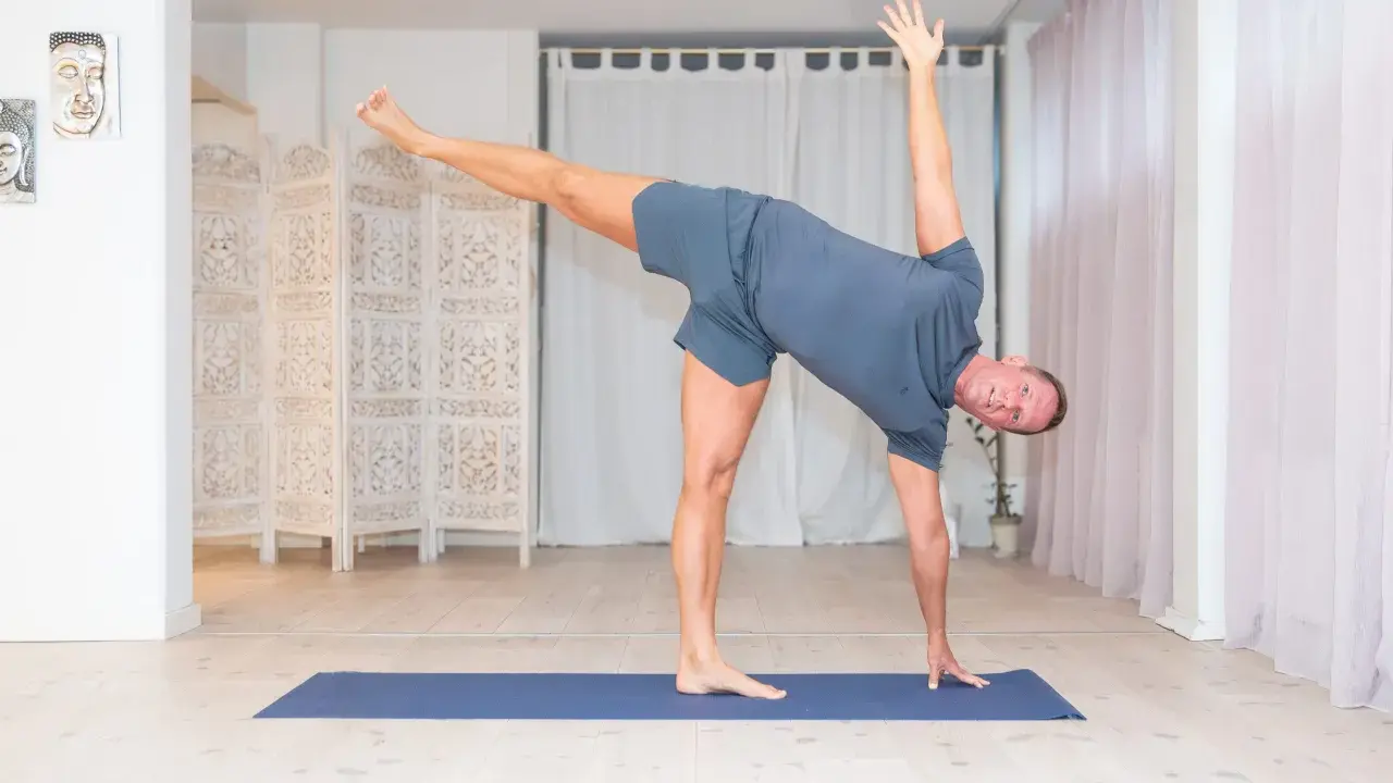 Man utför en dynamisk yoga-position på en blå matta i ett ljust rum med vita draperier och en vikbar rumsavdelare.