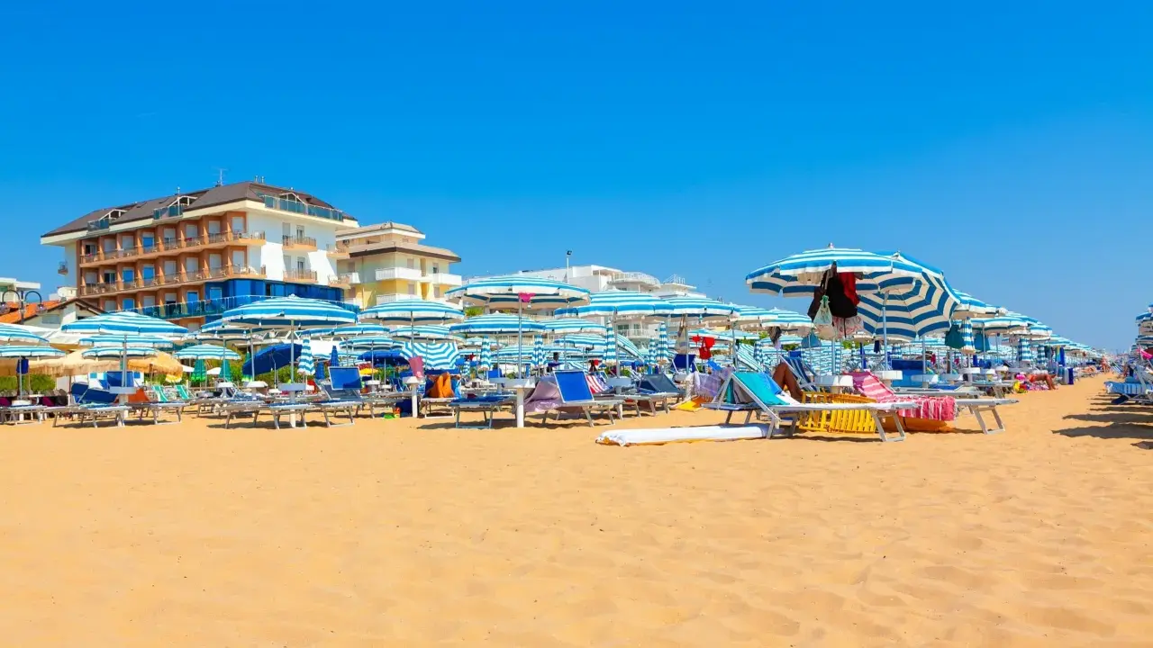 Słoneczny dzień na plaży w Lido di Jesolo. Rzędy leżaków i parasoli w niebiesko-białe pasy czekają na plażowiczów.