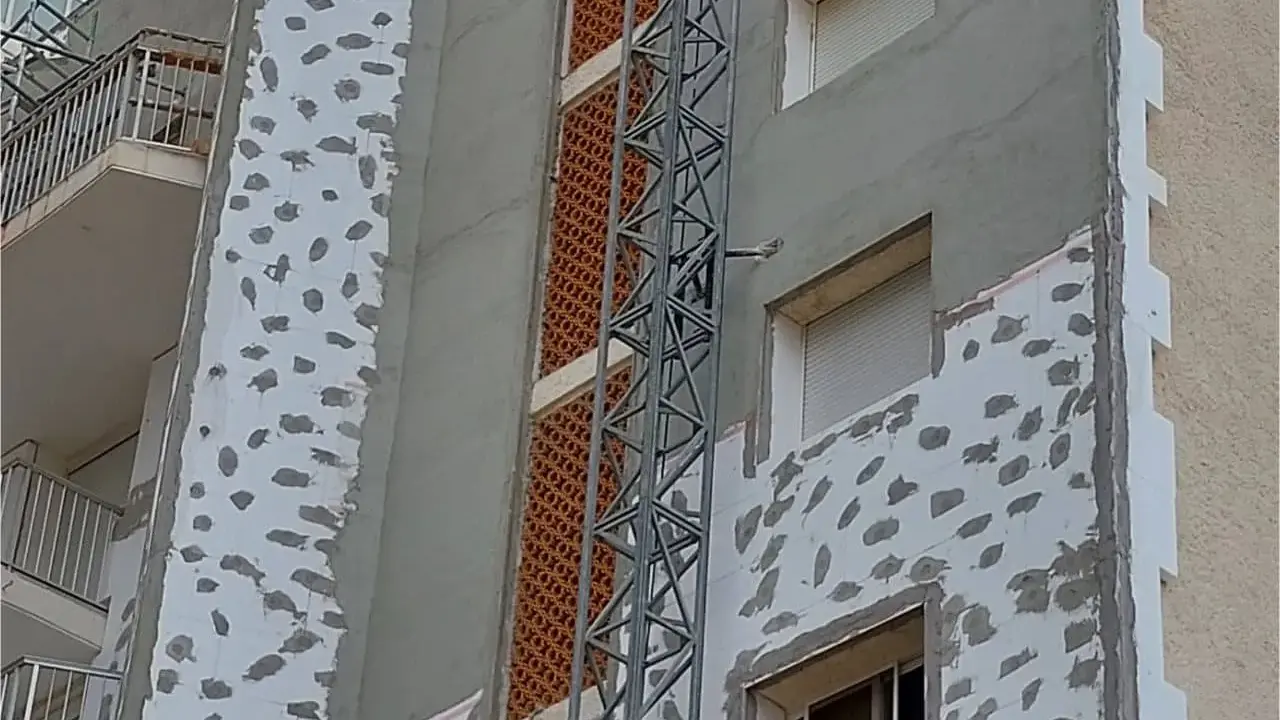 Proceso de como aislar una pared ya construida con placas de poliestireno y mortero. Se observa una grúa de obra.