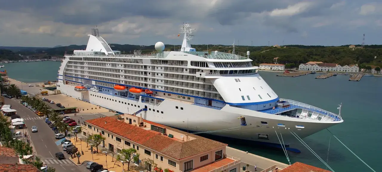 Grande nave da crociera attraccata nel porto Mahon, con edifici storici e colline verdi sullo sfondo.