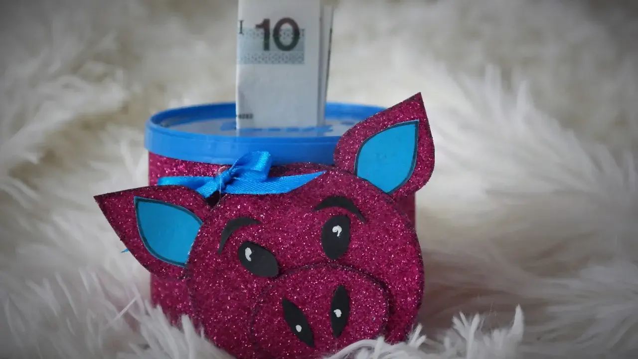 Urocza skarbonka diy w kształcie różowej świnki z brokatem, z niebieskimi uszami i kokardką. W środku widać banknot 10 zł.