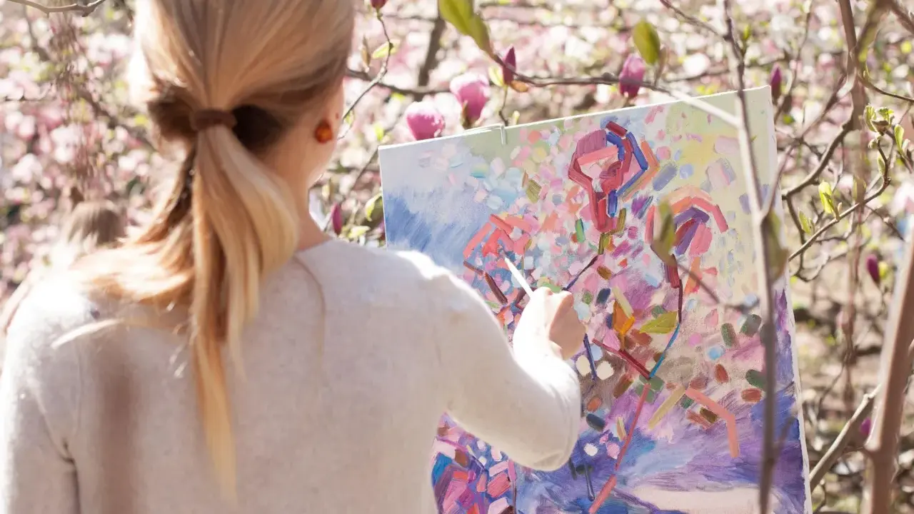 En kvinna målar en abstrakt tavla utomhus bland blommande magnoliaträd. Hon använder olika målartekniker för att fånga vårens färger.