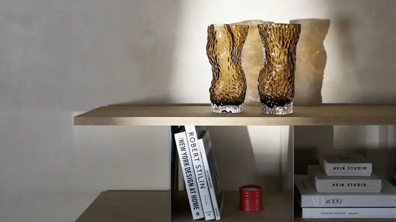 Två bärnstensfärgade glasvaser på en hylla, bredvid böcker och en röd ljushållare. Även böcker med texten HEIN STUDIO syns.