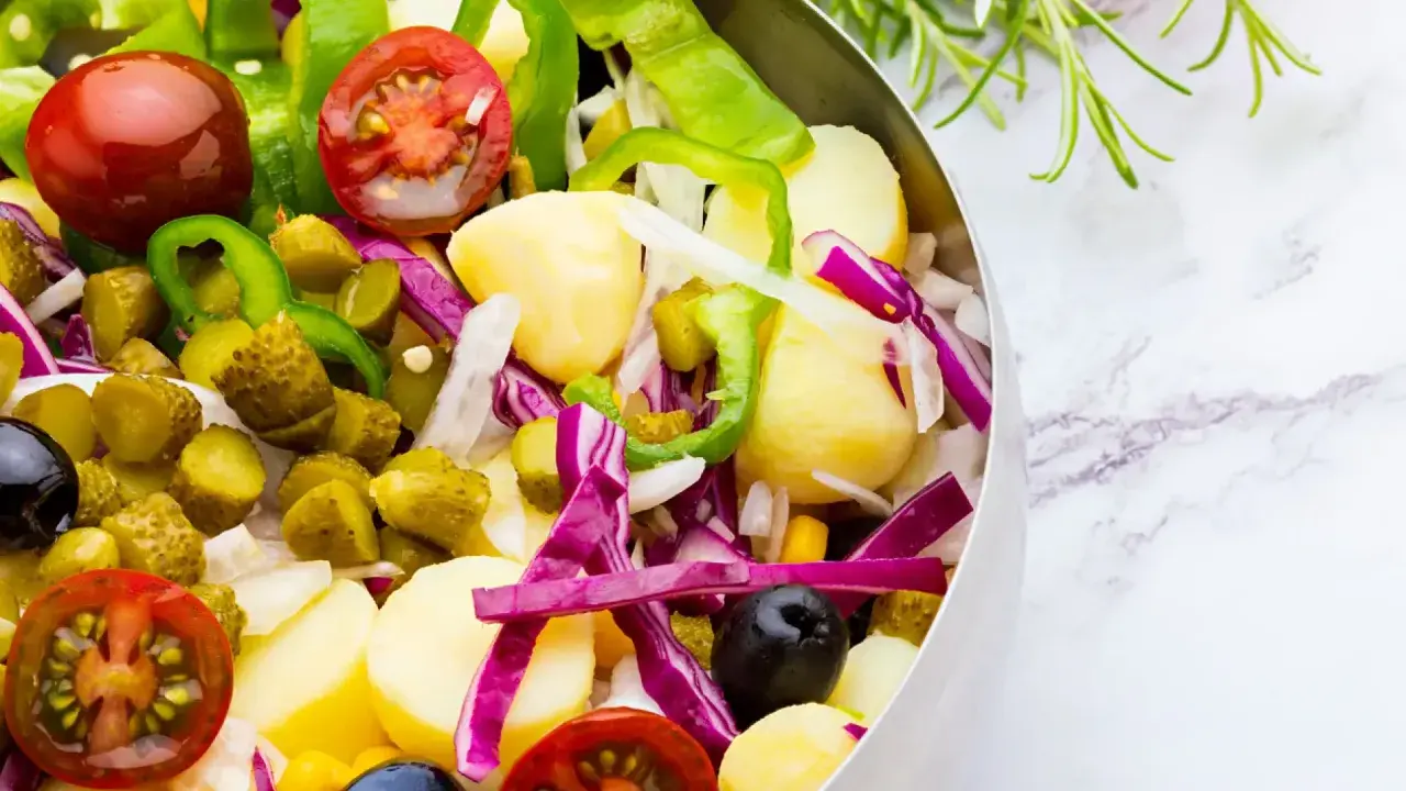 Ensalada campera vibrante con patatas, tomates, pepinillos, pimientos verdes, cebolla morada y aceitunas negras en un bol metálico.