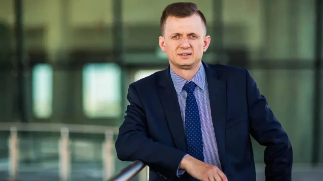 Mężczyzna w garniturze i krawacie opiera się o barierkę.