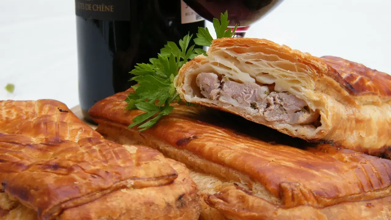 Pâté en croûte, une spécialité lorraine savoureuse, avec une garniture de viande visible. Une bouteille de vin rouge et du persil complètent la scène.