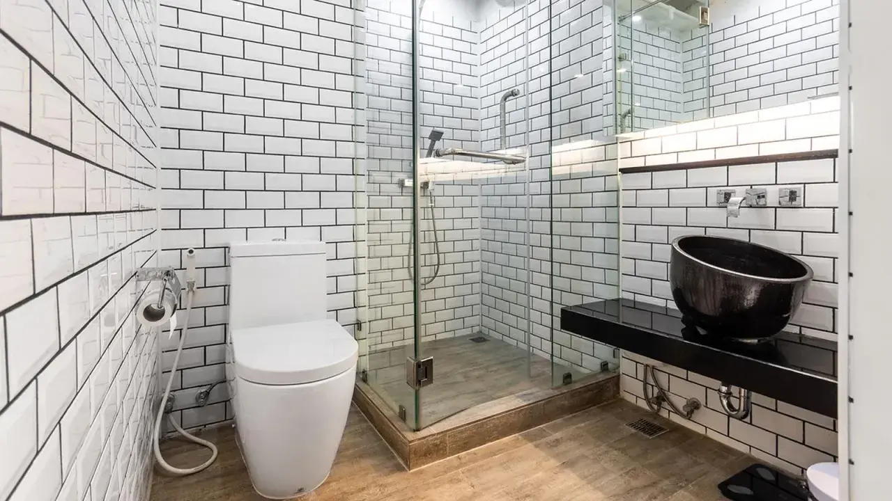Baño moderno con azulejos blancos y negros. El suelo de madera se ve primero, luego los azulejos. Inodoro, ducha de cristal y lavabo negro.