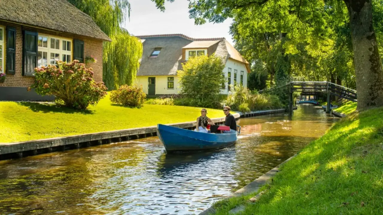Ein Paar fährt mit einem blauen Boot durch einen Kanal im "Klein Venedig Holland". Reetgedeckte Häuser und grüne Ufer säumen den Weg.