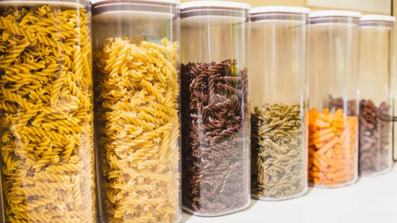 Contenitori trasparenti pieni di pasta a forma di farfalline, di vari colori: gialle, marroni, verdi e arancioni.