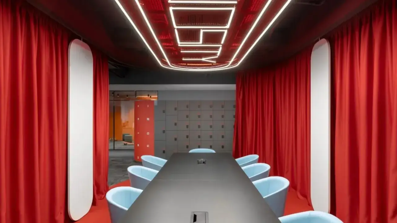 Aménagement salle de réunion moderne avec table noire, fauteuils bleus, rideaux rouges et éclairage néon au plafond.