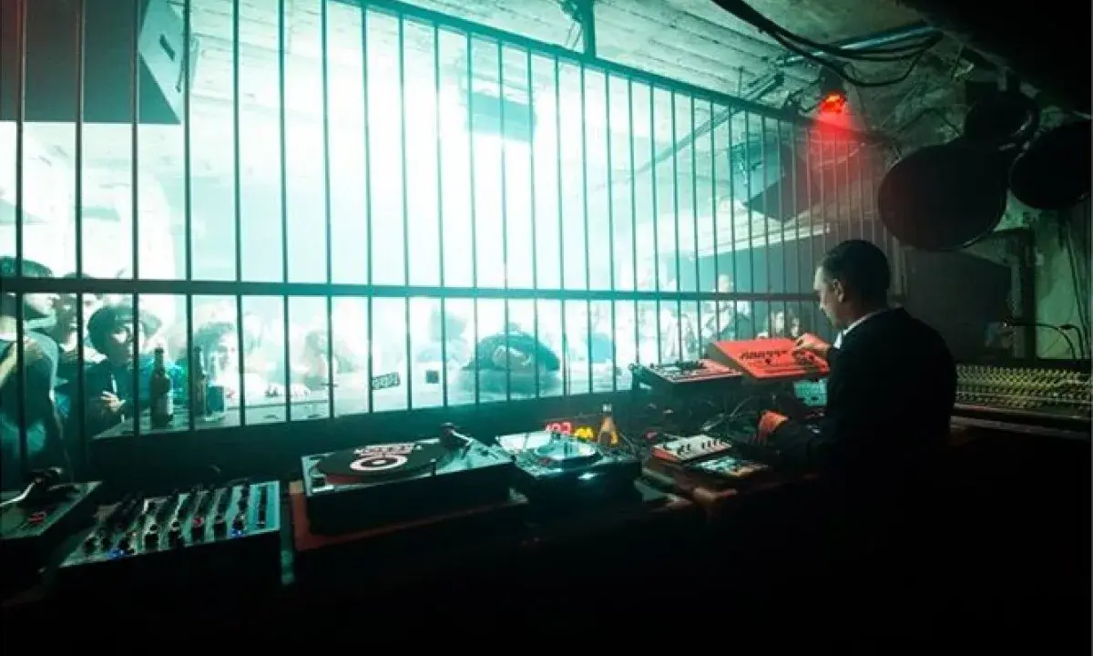 DJ set in un locale underground, luci al neon e folla che balla dietro le sbarre. Atmosfera da berlino rave.
