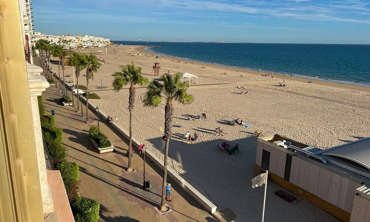 La **playa de la Costilla** con palmeras y gente disfrutando del sol. El mar azul se extiende hasta el horizonte.