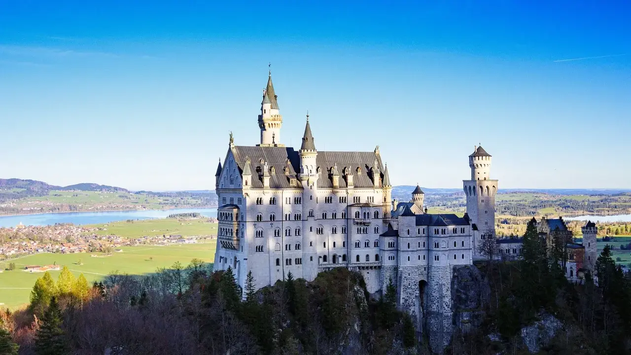 Sagolikt slott i Tyskland, Neuschwanstein, reser sig majestätiskt på en klippa med en sjö och grönskande landskap i bakgrunden.