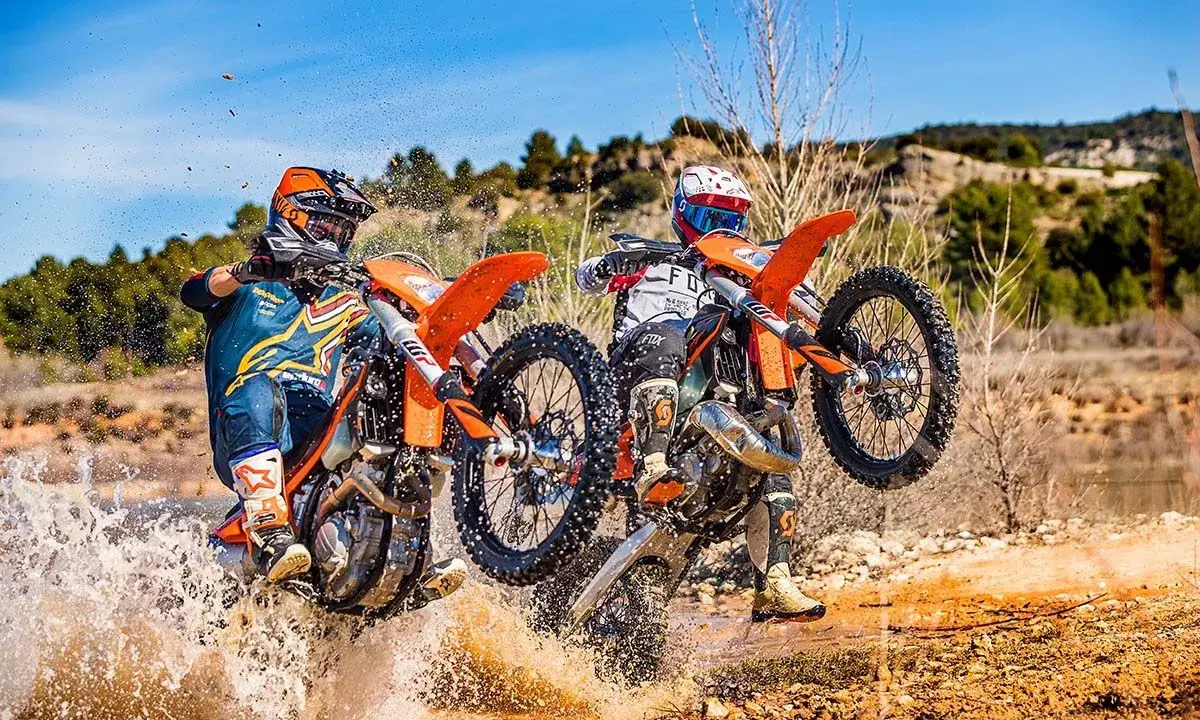 Dos pilotos de enduro compiten en un terreno embarrado, uno con motor 2t y otro 4t, salpicando agua.