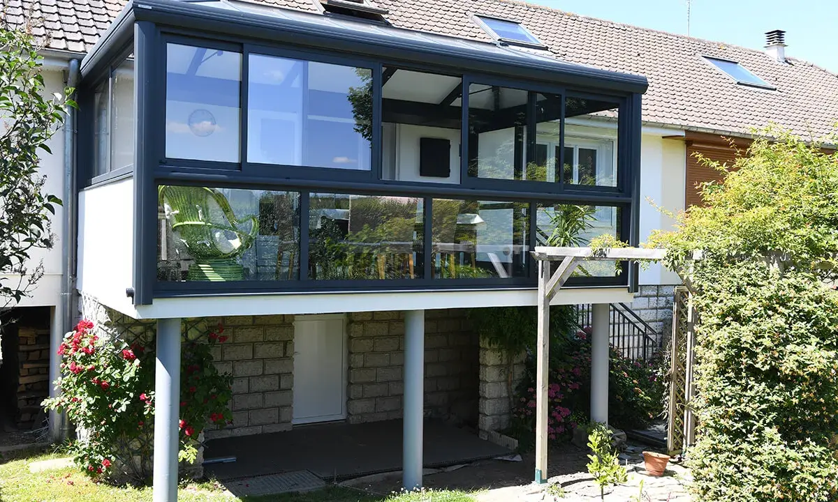Une extension véranda toit plat moderne, avec de larges baies vitrées, s'intègre parfaitement à la maison. Un fauteuil vert est visible à l'intérieur.