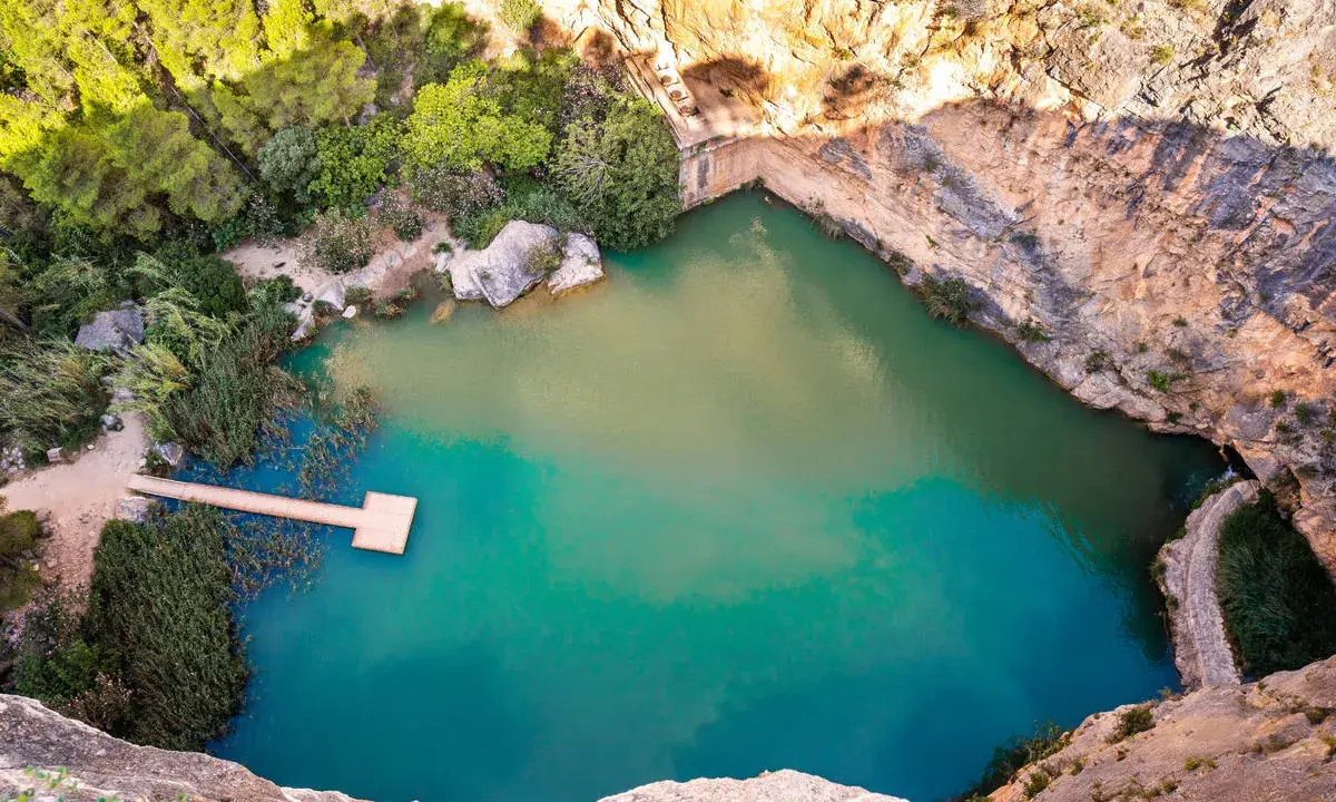 Un **charco azul** en Chiva, rodeado de rocas y vegetación. Un muelle de madera se adentra en sus aguas turquesas.