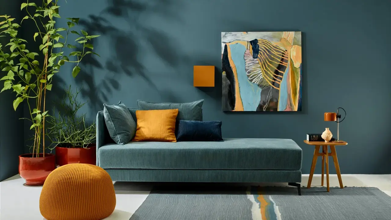 Ein Wohnzimmer mit einem türkisfarbenen Sofa, orangefarbenen Akzenten und einem abstrakten Gemälde. Die Wandfarbe könnte die Farbe des Jahres 2026 sein.